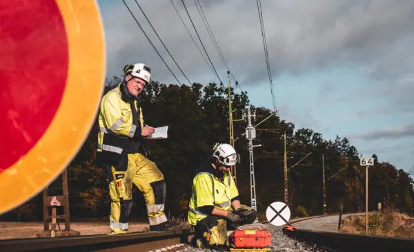 Två medarbetare på Strukton ute på arbete i spårmiljö, med varselkläder och vita hjälmar.